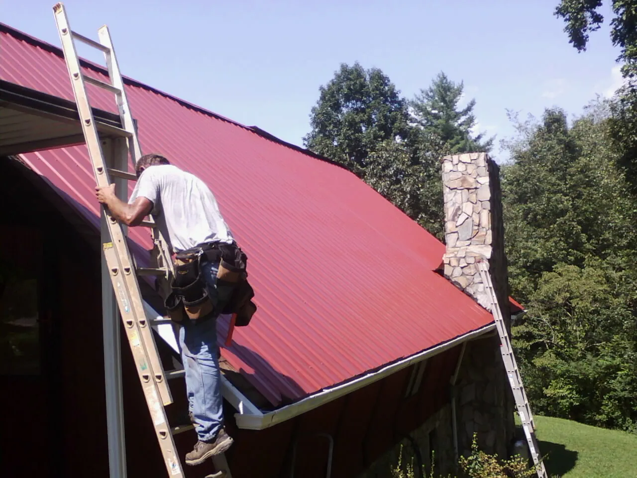 Roofer on a ladder beside a red metal roof and stone chimney — Soffit & Fascia Repair by Roofing Brothers in Glyndon
